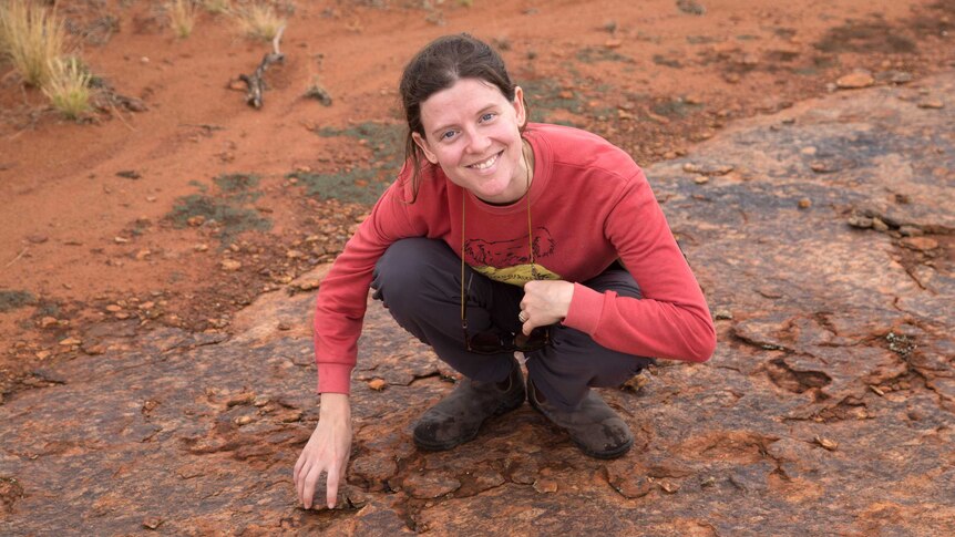Tamarah King poses for a photo smiling while kneeling down to the ground over historic earthquake damage in Central Australia.