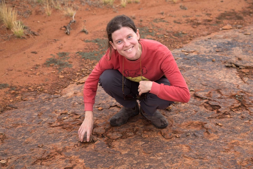 Tamarah King poses for a photo smiling while kneeling down to the ground over historic earthquake damage in Central Australia.