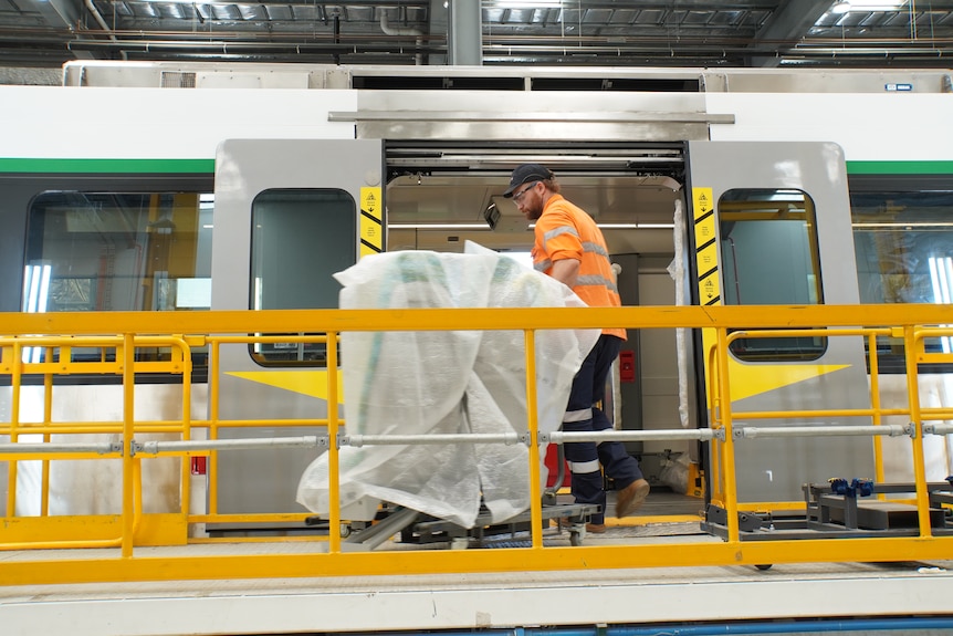 Workers in orange hi-vis clothing and safety gear, working on train parts at a railcar manufacturing facility.