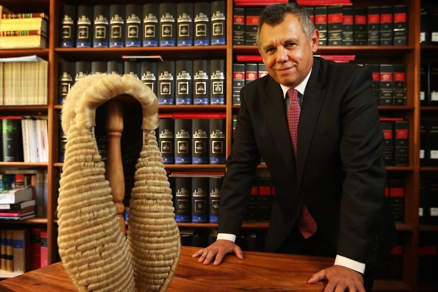 A man waring a suit is leaning on a desk that has a judges wig on it