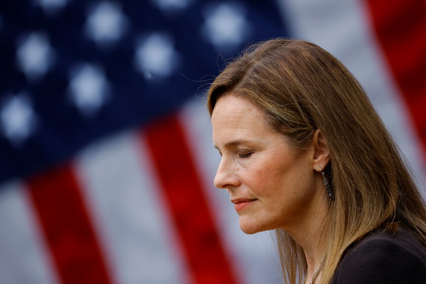 Amy Coney Barrett looks down with her eyes closed in front of a United States flag