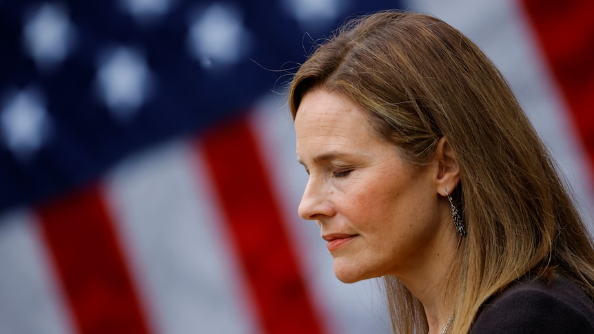 Amy Coney Barrett looks down with her eyes closed in front of a United States flag