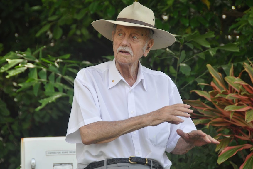 A man speaking to a crowd with a backdrop of tropical plantlife.