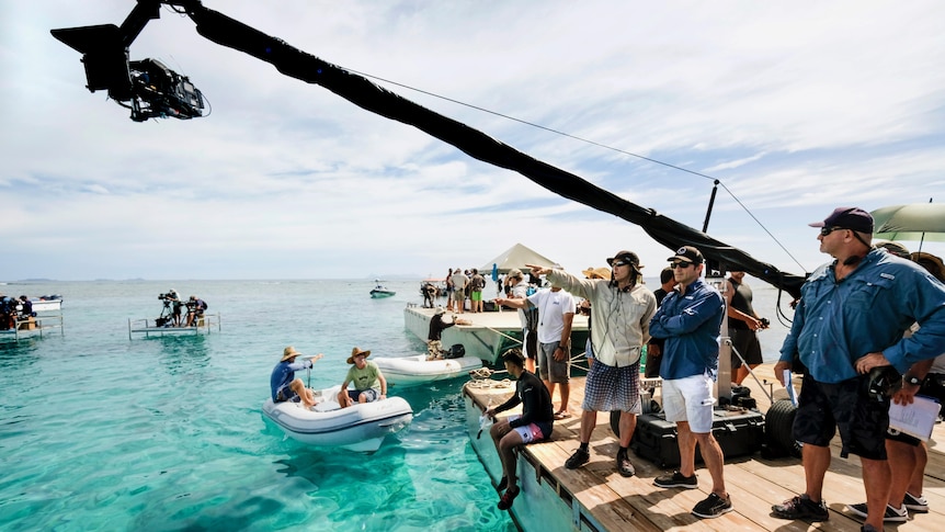 A film crew stands on a wooden boardwalk next to bright blue water. 