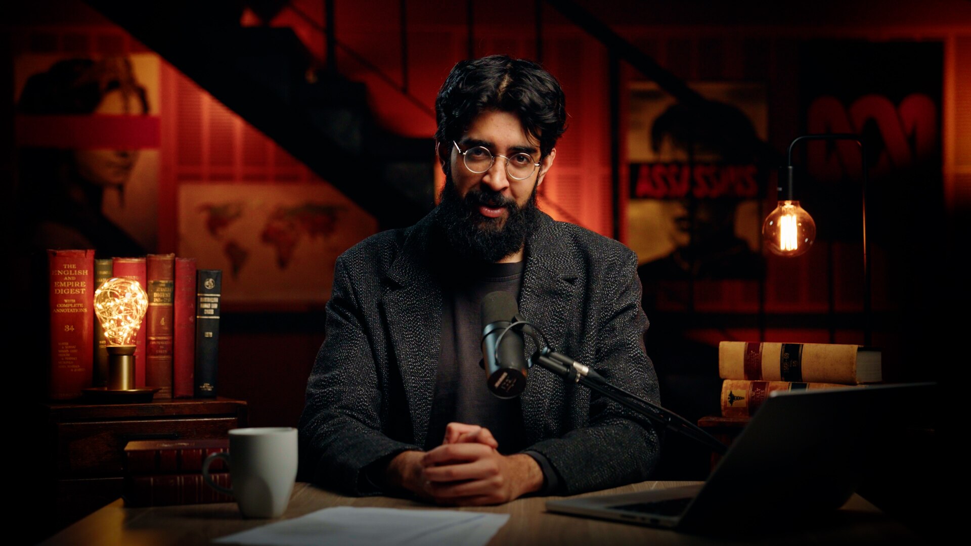 A person with dark hair and beard and sitting hands clasped in a dimly lit room 