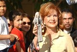 A red-headed woman wearing a yellow patterned top holds up a Barbie doll, with hands adorned with red nail polish