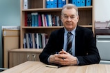 A man wearing a dark suit jacket and pale blue shirt sits at a desk in front of a bookshelf with his hands clasped.