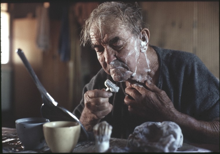 An older man looks in a mirror and shaves his face. 