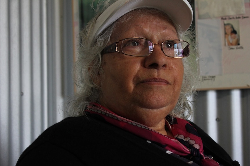 A clos up on the face of an older woman wearing glasses, pictured inside a structure made of corrugated iron