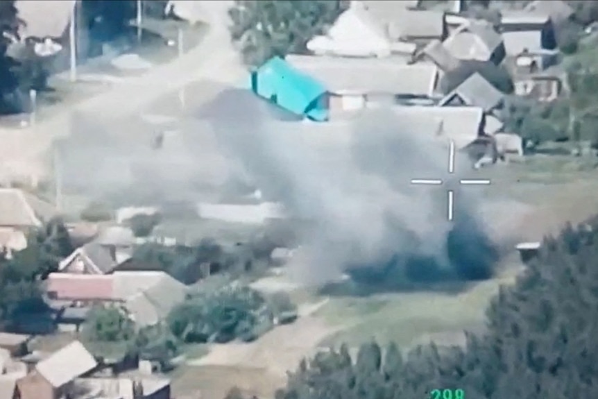 A still from a video taken from a drone. smoke rises over a green field dotted with buildings
