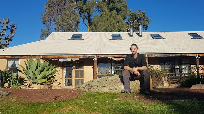 A smiling man sits on a rock in front of his rural home.