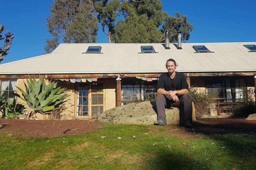 A smiling man sits on a rock in front of his rural home.