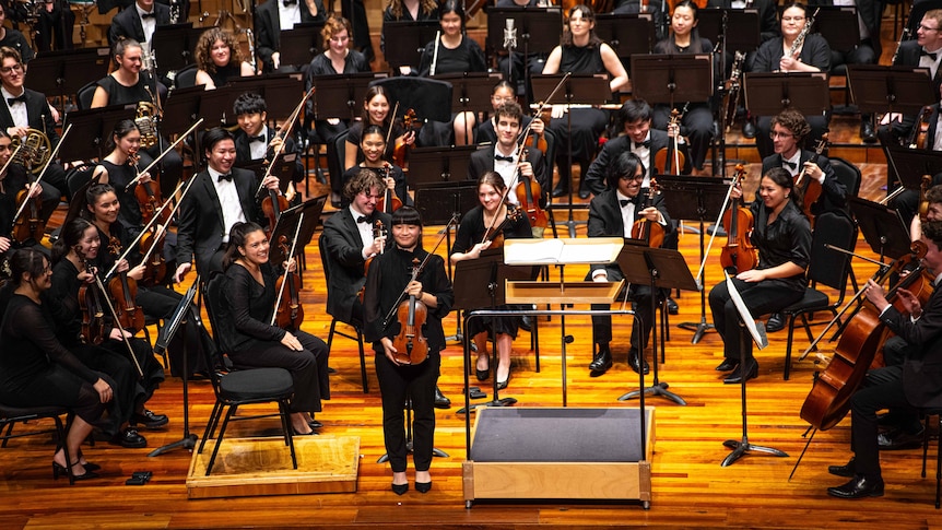 An orchestra of young musicians on stage. One violinist, the concertmaster stands facing the audience