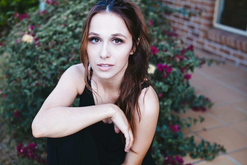 A young white woman poses in front of a bush with purple flowers