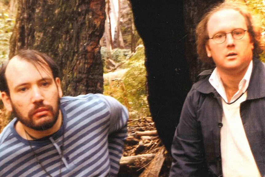 Two men look at a camera, forest in background.