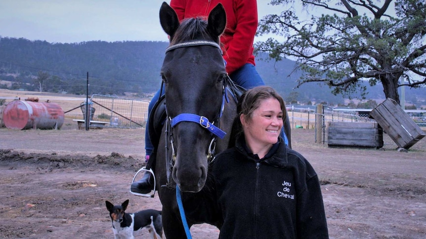 A woman stands next to a horse with a rider, holding the reins  on a rural property.