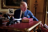 the king at a red leather topped desk, reaches out to a red box with a ribbon tied scroll, wearing blue pinstrip suit