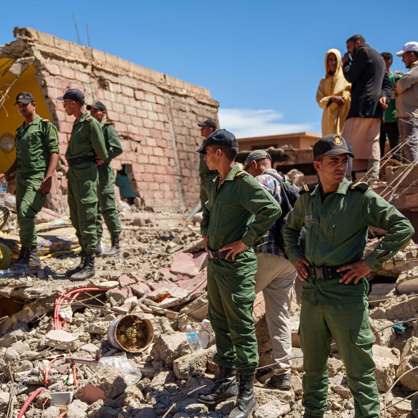 A group of people dressed in green uniform and wearing caps stand among rubble and look for people.
