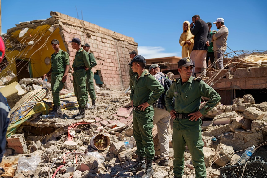 A group of people dressed in green uniform and wearing caps stand among rubble and look for people.