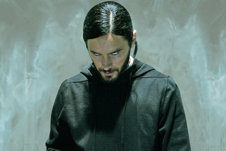 A pale man with dark hair and glowing eyes, wearing dark clothing, stands looking menacing in front of an icy background.