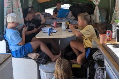 Four children sit around a table inside a cramped caravan, on phones and laptops. 