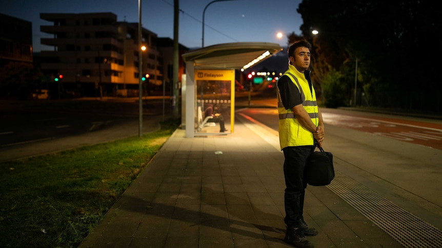 A man stands at a bus stop in the early morning.
