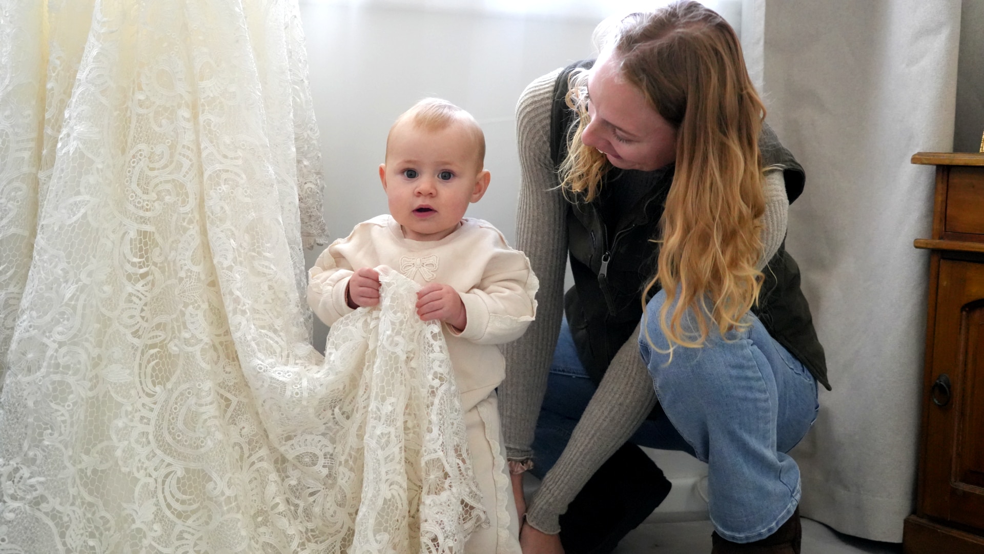 Baby wearing white standing holding the train of a lace wedding dress and looking at the camera with mum crouched beside