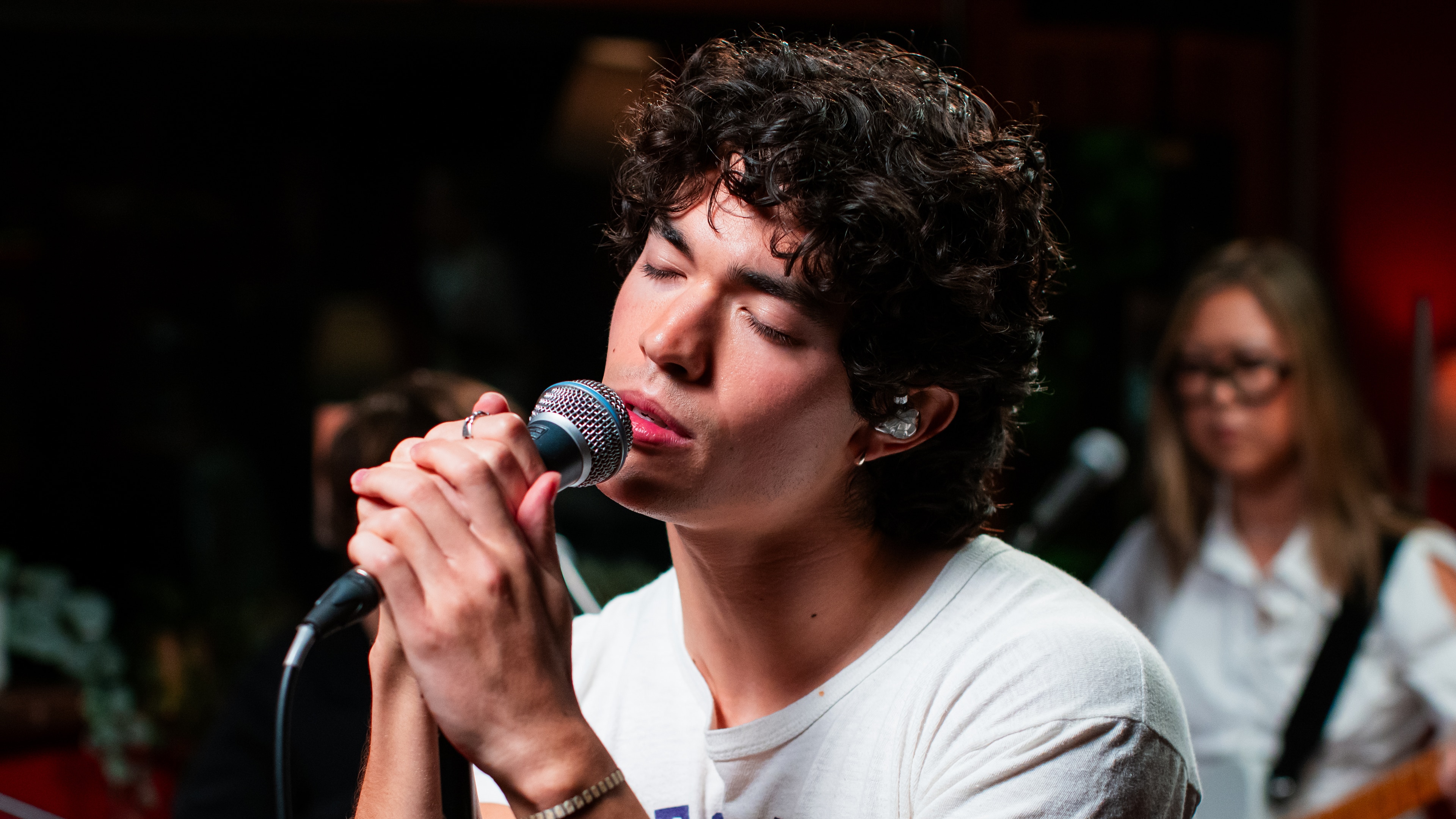 Conan Gray wears a white t shirt and sings into a microphone with his eyes closed.
