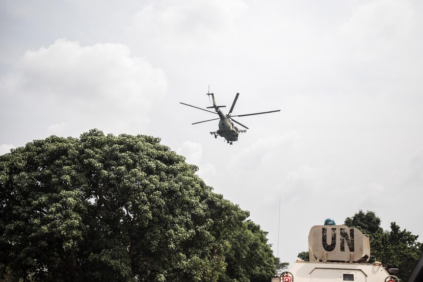 A helicopter hovers in the sky above the top of a vehicle with UN written on it 