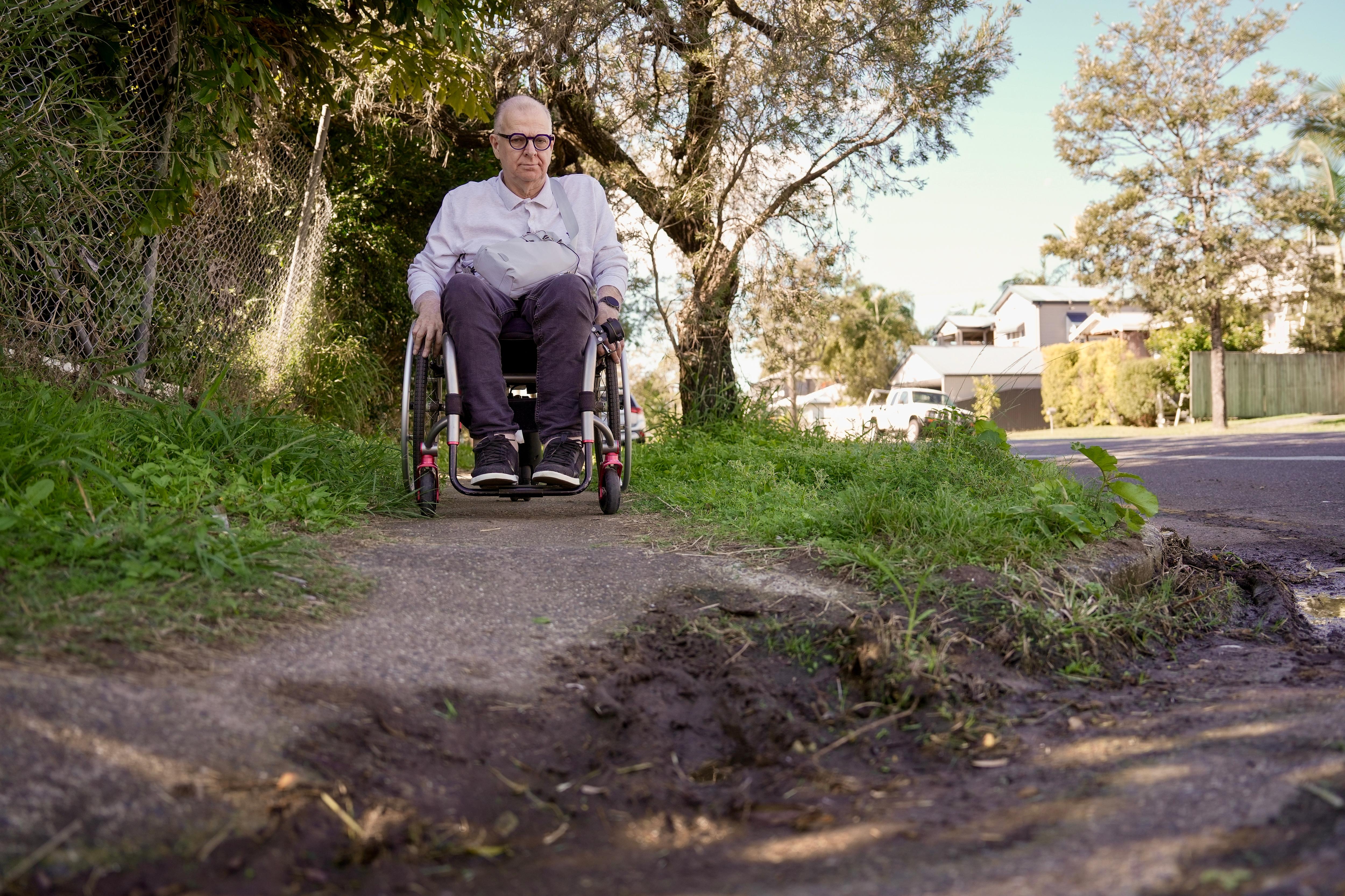 A white man with short grey hair. He's in a wheelchair looking at a muddy curb his chair won't be able to make it through