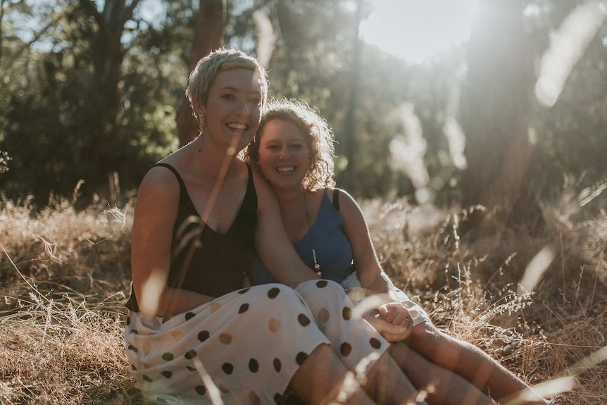 The couple, both blonde women in their late twenties, smile while holding hands and sitting in the afternoon sun.