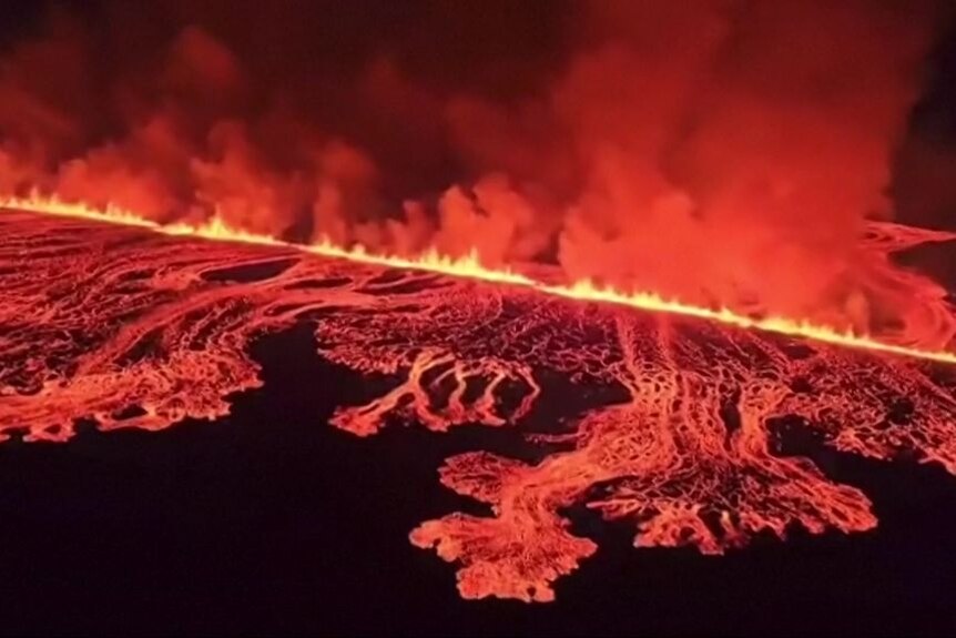 Aerial vision shows a red glow and lava flows with a wall of flames and plumes of smoke.