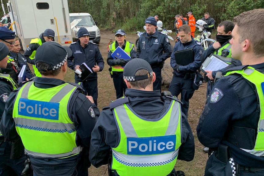 Police officers stand in a circle for a briefing in a dirt carpark in a state forest.
