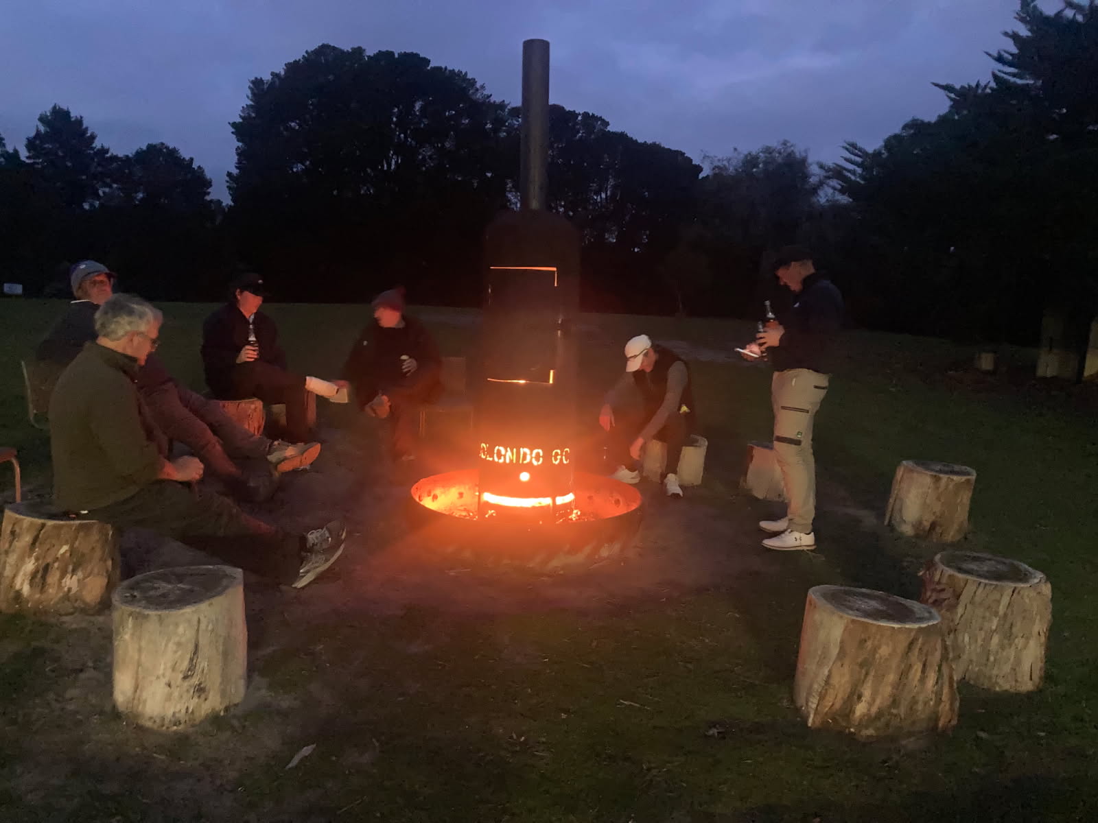 Six men sit on wood logs around a fire with beer in hand.
