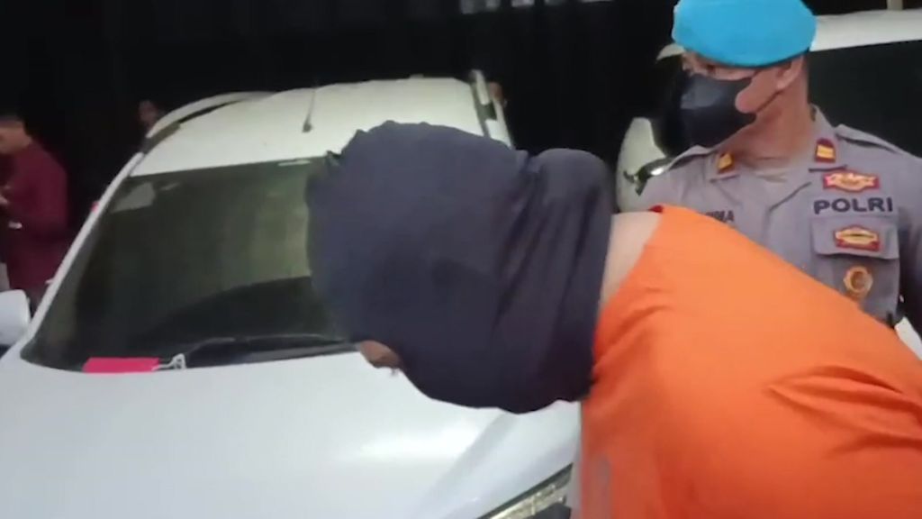 Un hombre que lleva un pasamontañas agacha la cabeza mientras un agente de policía lo conduce junto a un coche.