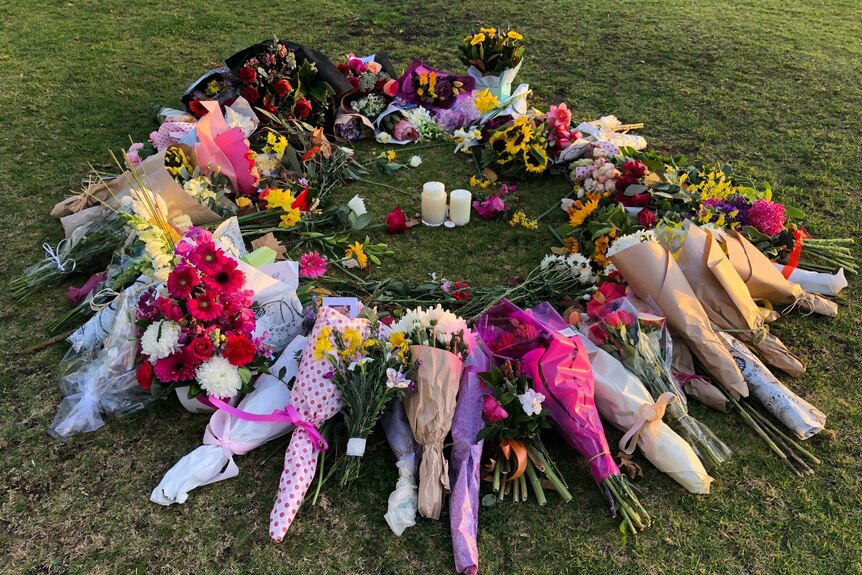 A circle of flowers and cards surround two candles on the Princes Park oval in afternoon sun.