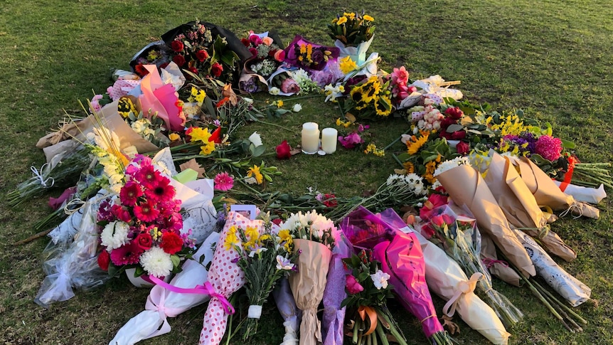 A circle of flowers and cards surround two candles on the Princes Park oval in afternoon sun.