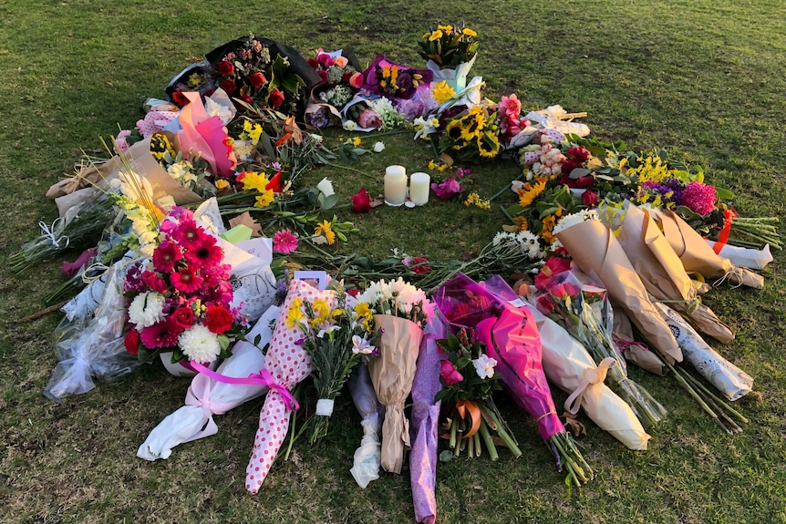 A circle of flowers and cards surround two candles on the Princes Park oval in afternoon sun.