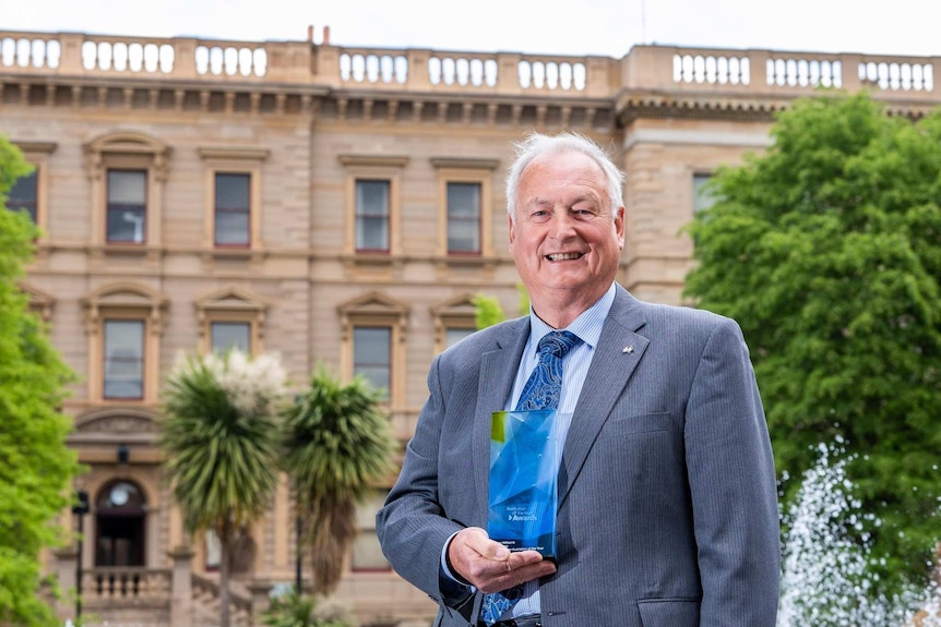 Man in suit smiling holding trophy standing in front of an old building with green leafy trees in background.