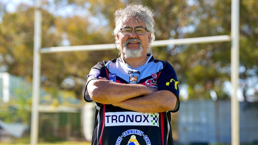 A man with curly grey and white hair smiles and crosses his arms on a football field.