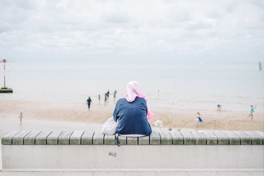 Woman wearing a pink hijab and appearing lonely while looking out over a beach on an overcast day to depict loneliness.
