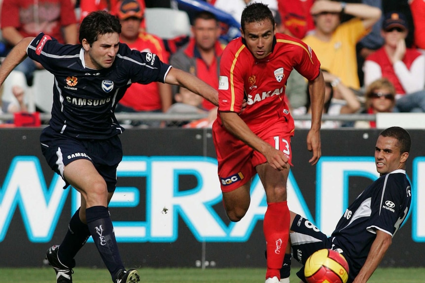 An A-League player races after the ball while a defender sits on the ground after missing a tackle.