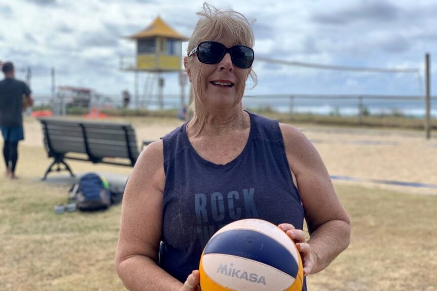 A woman wearing sunglasses hold a volleyball as she poses for a photograph in front of a beach volleyball court