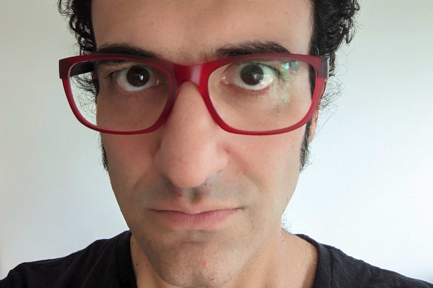 A man with curly hair and red glasses looks at the camera.