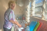 A woman with blonde hair washes dishes in a sink in her kitchen, near open louvered windows.