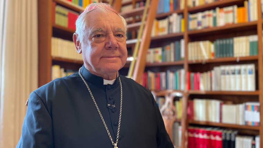 A man in black Catholic robes and wearing a cross around his neck looks at the camera
