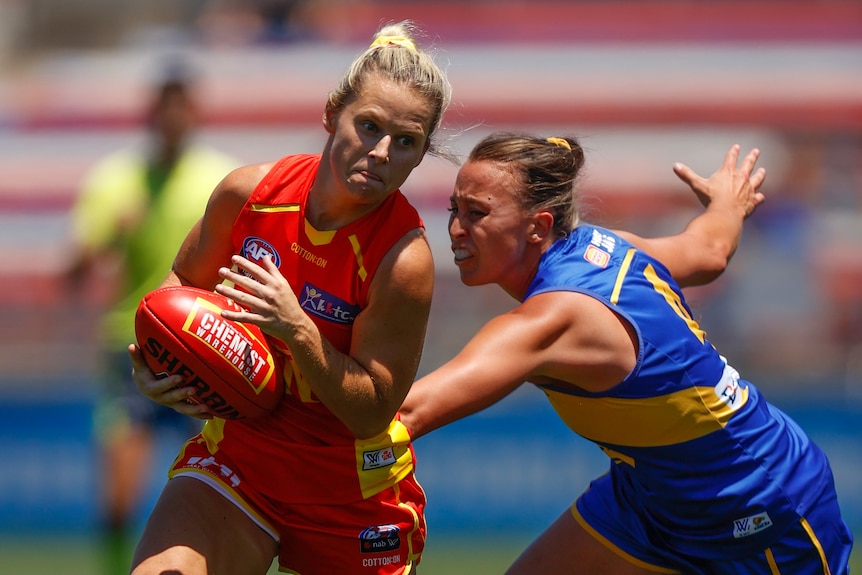 A Gold Coast Suns AFLW player runs with the ball as she beats the tackle of a West Coast opponent.