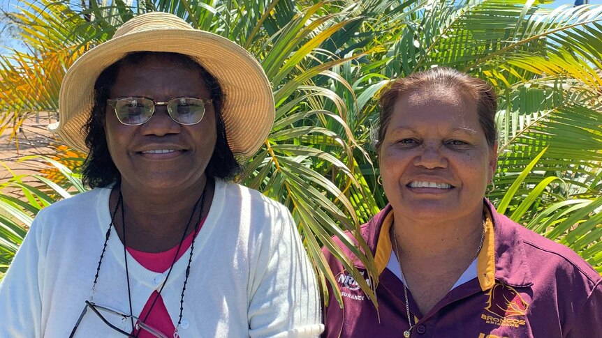 Anita Painter, left, wears a hat next to Helen Lee, right, as they stand in front of palm trees with blue skies overhead.