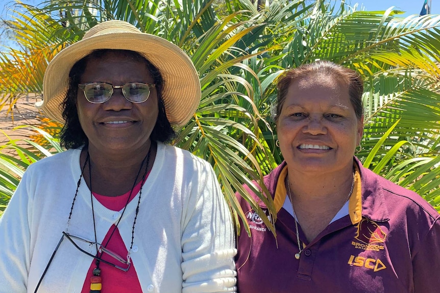 Anita Painter, left, wears a hat next to Helen Lee, right, as they stand in front of palm trees with blue skies overhead.