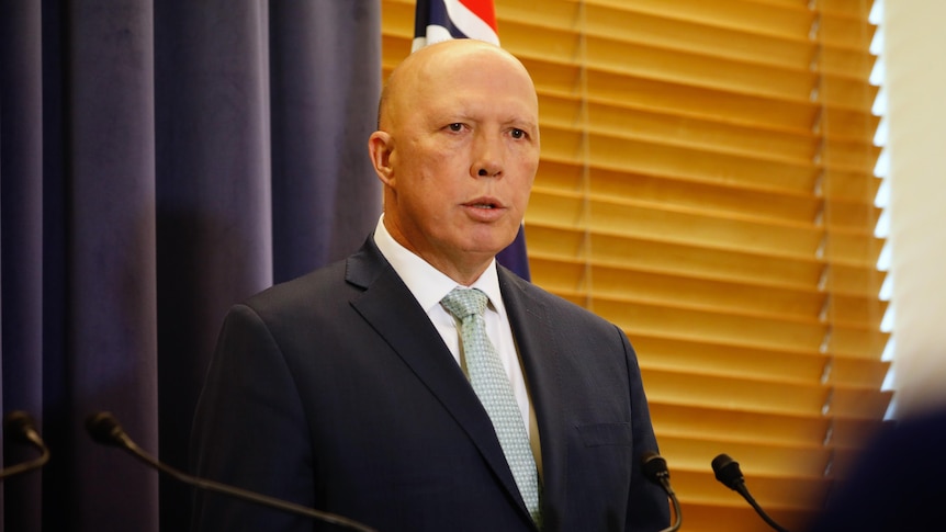 A bald man in a suit stands in front of an Australian flag, a blue curtain and a wooden-slatted window.